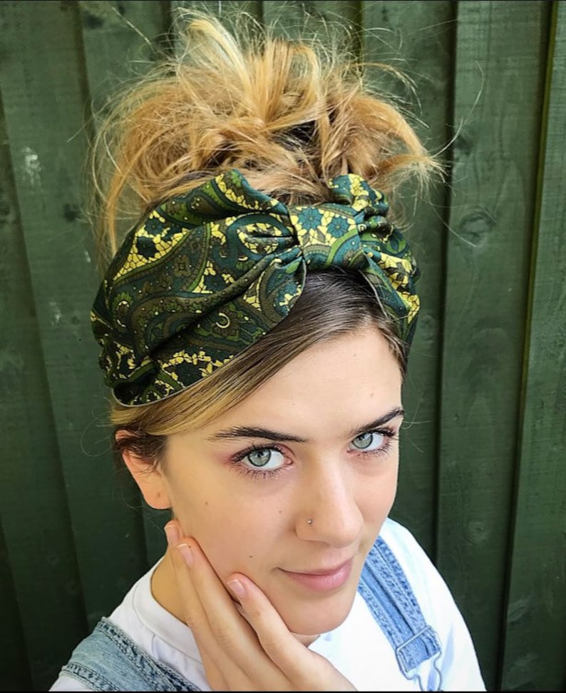 May include: A green and gold paisley patterned headband. The headband is tied in a knot on top of the head. The person in the image is wearing a white t-shirt and denim overalls.