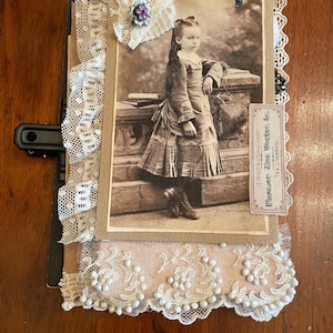 May include: Vintage scrapbook featuring a sepia-toned portrait of a young girl in a dress, accented with lace trim and a decorative flower. The scrapbook includes a label with text. The overall design is antique and nostalgic.