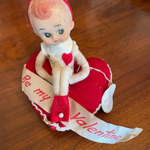 May include: Vintage Valentine's Day decoration featuring a doll with a red cap and red shoes, seated on a red heart. A white ribbon with the words "Be my Valentine" is draped across the heart. The doll has a white outfit and a red heart on its chest.