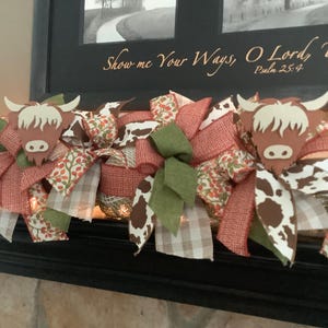 May include: A decorative garland featuring Highland cow cutouts, red, green, and brown ribbons, and cow print accents. The garland is adorned with a variety of patterned ribbons and bows, creating a rustic and charming aesthetic. The garland is placed on a black surface.