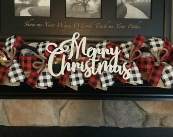 May include: A Christmas garland with a "Merry Christmas" sign. The garland features red and black buffalo check ribbon, burlap, and string lights. The background includes a framed picture and text that reads "Show me Your Ways, O Lord, Teach me Your Paths."