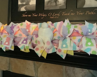 May include: Easter-themed decorative garland featuring pastel-colored ribbons with polka dots, stripes, and bunny designs. A plush white bunny with pink foot pads is centered. The garland is displayed on a black surface, with text above that reads "Show me Your Ways, O Lord, Teach me Your Paths."