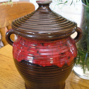 May include: A brown ceramic jar with a red glaze and a lid. The jar has a ribbed design and is decorated with a red and black pattern.