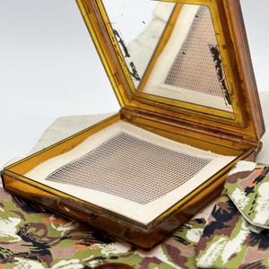 May include: Open, vintage amber-colored compact with a mirror and a mesh powder screen. The compact is rectangular with a hinged lid. A brown, round object and a green, brown, and white patterned fabric are in the foreground.