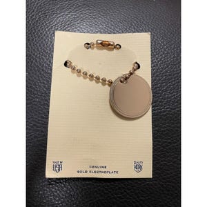 以下が含まれることがあります： 丸いタグとボールチェーンが付いた金メッキのキーホルダー。タグは空白で、チェーンにはロブスタークラスプが付いています。キーホルダーの裏のカードには、「Genuine Gold Electroplate」と「Made in USA」と書かれています。