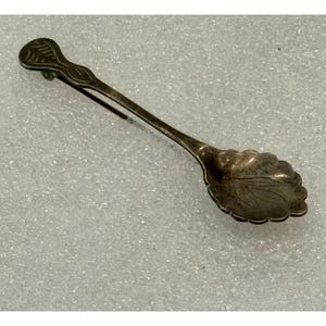May include: A silver-toned metal brooch shaped like a small spoon with a scalloped edge bowl and a long, slender handle. The handle features an engraved leaf-like pattern, and the bowl of the spoon is decorated with a subtle, etched leaf design.