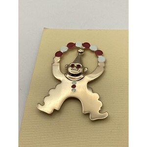 以下が含まれることがあります： 赤と白のボールをジャグリングするピエロの金色の金属製のピン。ピエロは赤い帽子をかぶり、赤い目をしています。