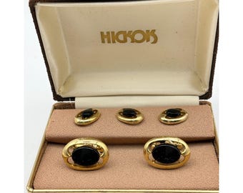 May include: Vintage gold-tone cufflinks and shirt studs set with black oval insets, presented in a brown velvet-lined box. The box is open, revealing the cufflinks and studs. The brand name "HICKOKS" is printed in gold on the inside of the box.