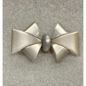 Könnte beinhalten: Eine silberfarbene Brosche in Form einer Schleife. Die Brosche hat einen zentralen, oval geformten, perlenartigen Akzent. Das Design der Schleife weist gefaltete, geometrische Formen auf, die einen geschichteten Effekt erzeugen. Die Brosche ist wahrscheinlich aus Metall.