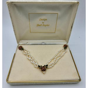 May include: A pearl necklace with three strands, featuring red rose accents with green leaves and a gold-colored box. The box has the text "Design by Shell Kepler" in gold lettering.