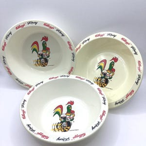 Op de afbeelding: Drie crèmekleurige keramische kommen. Elke kom heeft een cartoonhaan met een racehelm en de tekst "Kellogg's Racing" in het rood rond de rand. De haan is groen, rood, geel en zwart.