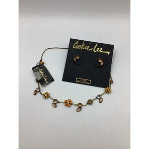 Pode incluir: Um colar de ouro com um design floral com cristais laranja e transparentes. O colar é exibido com brincos combinando em um cartão preto com o texto "Cookee Lee" em letras douradas.