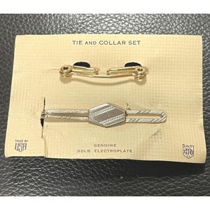以下が含まれることがあります： 紙製の箱に入った金のネクタイと襟のセット。箱には「Tie and Collar Set」、「Genuine Gold Electroplate」、「Made in USA」、「Quality Co.」と記載されています