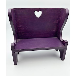 May include: A small, purple wooden bench with a heart-shaped cutout in the backrest. The bench has a simple design with a seat and four legs. The wood grain is visible, giving it a rustic appearance. The bench is painted in a solid, deep purple color.