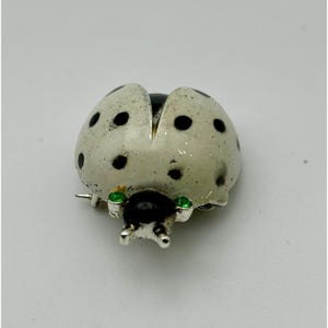 May include: A ladybug brooch with a white body speckled with black spots. The ladybug's head is black, and it has two small green gems for eyes. The brooch is made of metal.