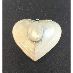 Op de afbeelding: Een hartvormige hanger met een parelmoer afwerking. Een ovaalvormige parel is in een zilveren montuur aan de voorkant gezet. De hanger heeft een kleine lus aan de bovenkant voor een ketting. Het hart is ongeveer 5 cm breed.