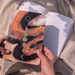 May include: A planner with a cover featuring a black and orange abstract swirl design and a vertical row of gold foil moon phases. The planner is open to reveal white pages with lined sections and small circular icons for tracking lunar cycles.