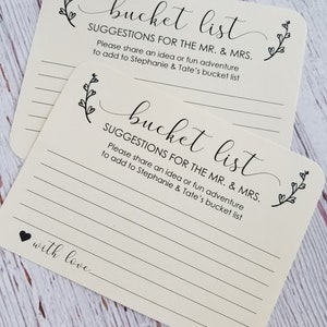 Op de afbeelding: Twee witte kaarten met zwarte tekst en bloemmotieven. De tekst op de kaarten luidt "bucket list" en "SUGGESTIES VOOR DE MAN EN DE VROUW. Deel een idee of een leuk avontuur om toe te voegen aan de bucketlist van Stephanie en Tate".
