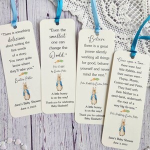 May include: Four white bookmark favors with Peter Rabbit illustrations and inspirational quotes. The bookmarks are tied with blue and light blue ribbon. The text on the bookmarks includes "Even the smallest one can change the world." "Believe there is a great power silently working all things for good, behave yourself and never mind the rest." "There is something delicious about writing the first words of a story. You never quite know where they'll take you." and "Once upon a time... there were four little Rabbits, and their names were - Flopsy, Mopsy. Cotton-tail and Peter. They lived with their Mother in a sand-bank, underneath the root of a very big fir-tree."