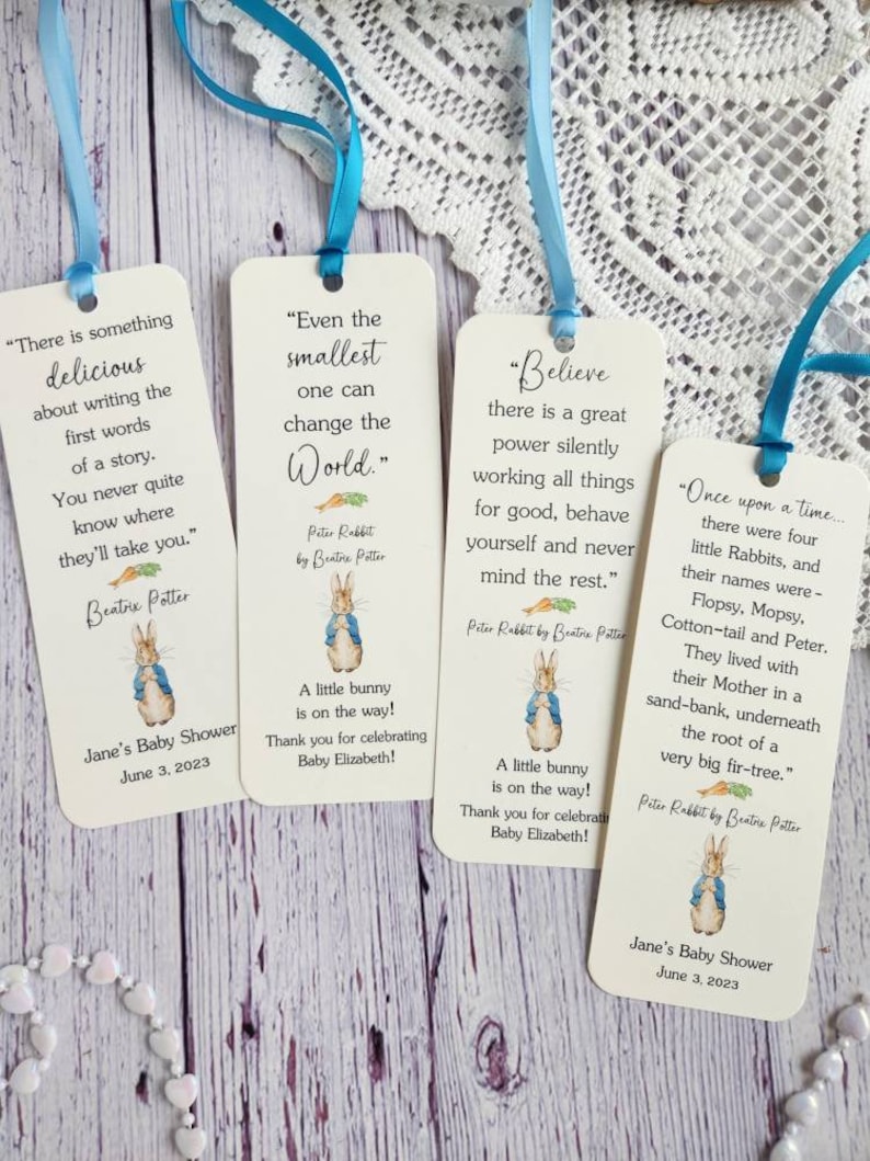 May include: Four white bookmark favors with Peter Rabbit illustrations and inspirational quotes. The bookmarks are tied with blue and light blue ribbon. The text on the bookmarks includes "Even the smallest one can change the world." "Believe there is a great power silently working all things for good, behave yourself and never mind the rest." "There is something delicious about writing the first words of a story. You never quite know where they'll take you." and "Once upon a time... there were four little Rabbits, and their names were - Flopsy, Mopsy. Cotton-tail and Peter. They lived with their Mother in a sand-bank, underneath the root of a very big fir-tree."