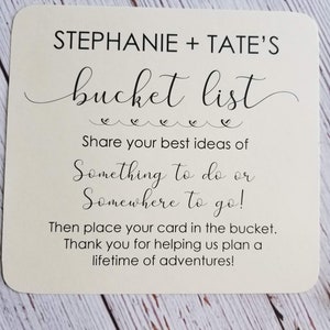 Op de afbeelding: Een witte kaart met zwarte tekst die "STEPHANIE + TATE'S bucket list" en "Deel je beste idee&euml;n voor iets om te doen of een plek om naartoe te gaan! Plaats je kaart dan in de emmer. Bedankt dat je ons helpt om een leven vol avonturen te plannen!" zegt.