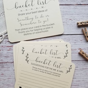 Op de afbeelding: Een set cr&egrave;mekleurige kaarten met zwarte tekst die "Amber & Derek's bucket list" luidt. De kaarten zijn bedoeld voor gasten om idee&euml;n voor de bucketlist van het stel op te schrijven.