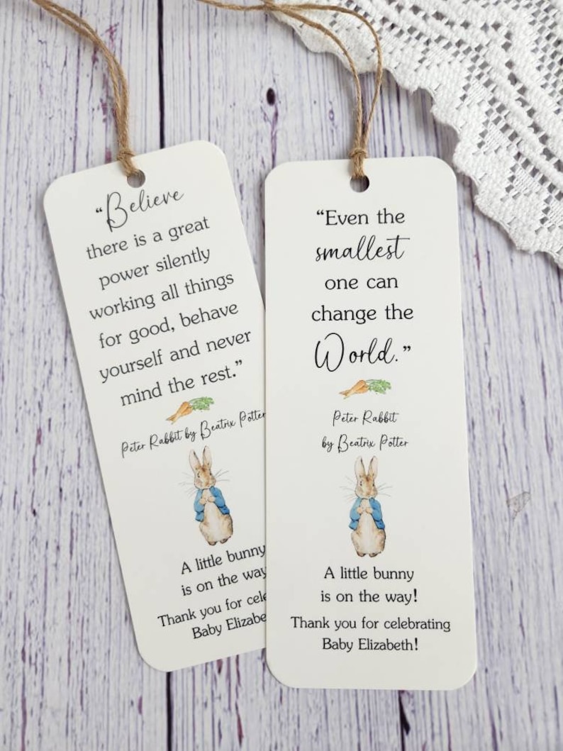 May include: Two white bookmark favors featuring Peter Rabbit illustrations and inspirational quotes. The first bookmark says "Believe there is a great power silently working all things for good, behave yourself and never mind the rest." The second bookmark says "Even the smallest one can change the World." Both bookmarks include the text "Peter Rabbit by Beatrix Potter" and "A little bunny is on the way! Thank you for celebrating Baby Elizabeth!"