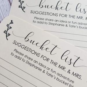Op de afbeelding: Twee witte kaarten met zwarte tekst en een eenvoudig bloemenmotief. De tekst op de kaarten luidt "bucket list" en "SUGGESTIES VOOR DE MAN EN DE VROUW. Deel een idee of een leuk avontuur om toe te voegen aan de bucketlist van Stephanie en Tate."