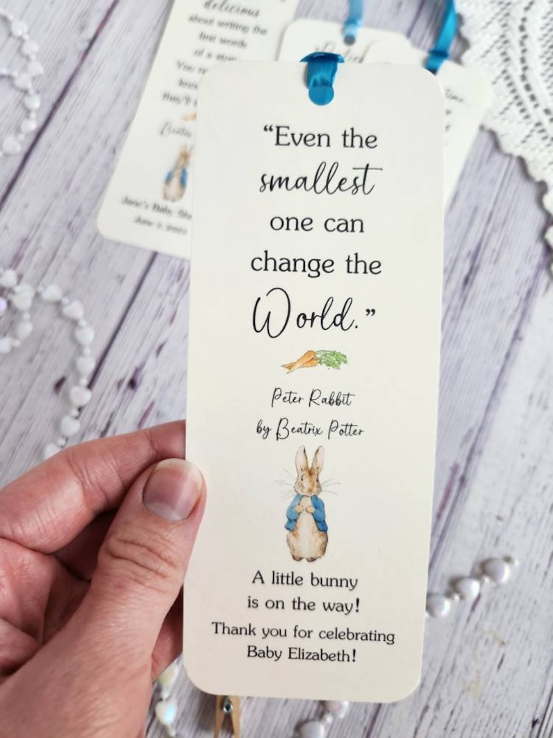 May include: A white bookmark with a blue ribbon. The bookmark features an illustration of Peter Rabbit from the book by Beatrix Potter. The text on the bookmark reads "Even the smallest one can change the world." and "A little bunny is on the way! Thank you for celebrating Baby Elizabeth!"