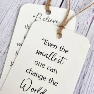 May include: Two white bookmark tags with inspirational quotes. The top tag says "Believe" and the bottom tag says "Even the smallest one can change the world." with a small illustration of carrots and parsley.