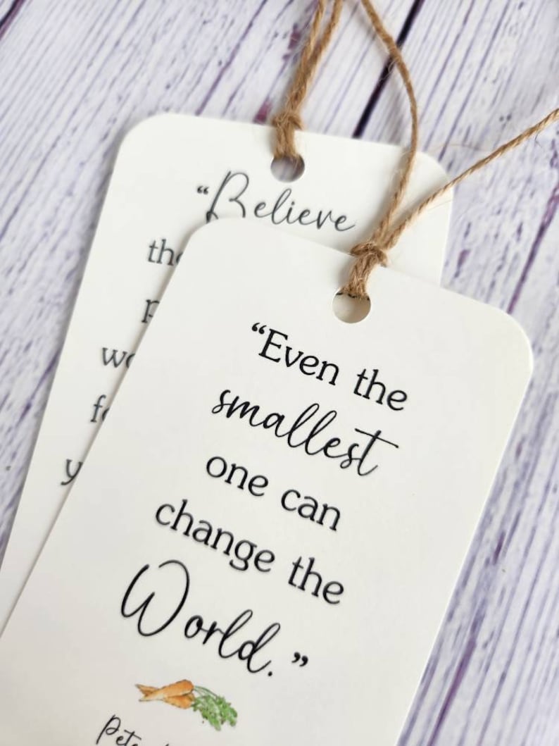 May include: Two white bookmark tags with inspirational quotes. The top tag says "Believe" and the bottom tag says "Even the smallest one can change the world." with a small illustration of carrots and parsley.