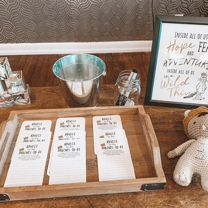 May include: A wooden tray containing eight white cards with the inscription "Advice for the Parents-to-Be". There is a silver bucket, a glass jar with black markers, and a crocheted stuffed toy with a crown on its head. A sign above the tray reads "Inside all of us is a Wild Thing".