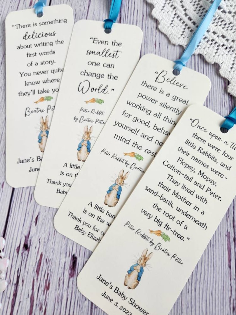 May include: Four white bookmarks with blue ribbon ties feature illustrations of Peter Rabbit from the children's book by Beatrix Potter. Each bookmark has a different quote from the book. The bookmarks are perfect for a baby shower or birthday party.
