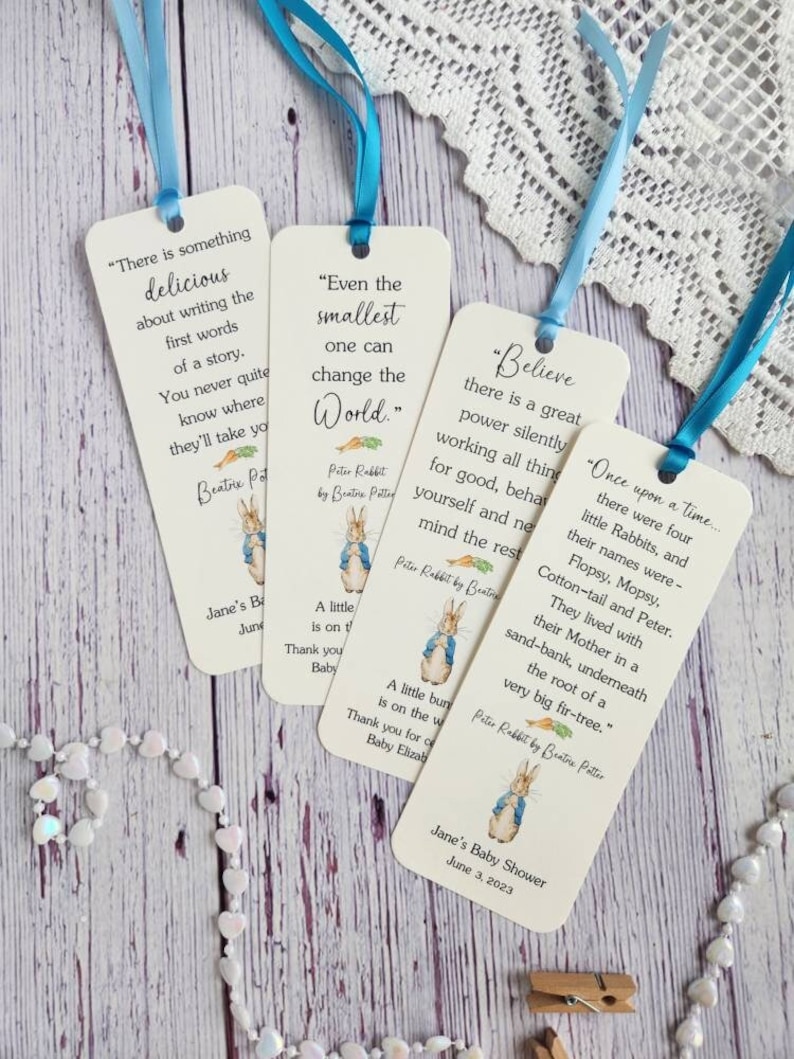May include: Four white bookmark favors featuring Peter Rabbit illustrations and quotes from the book. The bookmarks are tied with blue ribbon and have the text "Jane's Baby Shower June 3, 2023" printed on the bottom.