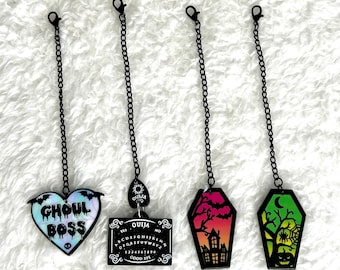 May include: Four black chains with different Halloween-themed charms. The charms include a heart with the text "Ghoul Boss", a Ouija board, a haunted house, and a coffin with a spider and a crescent moon.