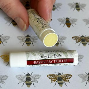 May include: A white tube of lip balm with a green and white label that reads "All Natural Bath & Beauty Products Silky Lip Balm Raspberry Truffle  Paraben Free Gluten Free Nut Free Vegetarian Piper Dee Naturals".