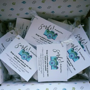 May include: An open box filled with white tags featuring a teal camera graphic and the word "Smile!". The tags include instructions to track results. The box has a patterned interior.