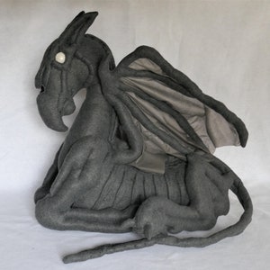 Może przedstawiać: Szary pluszowy smok z długim ogonem i dużymi skrzydłami. Smok ma białe oko i szczegółową, fakturowaną powierzchnię.