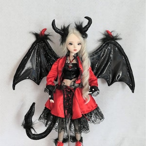 May include: A doll dressed in a red satin outfit with black lace accents, complete with black wings, horns, and a tail. The doll has long, flowing blonde hair and is wearing red boots. The overall theme is gothic fantasy.