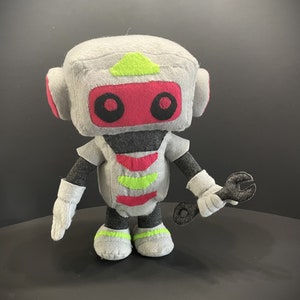 May include: A gray and red robot plush toy with a green triangle on its head. The robot is holding a black wrench in its right hand.