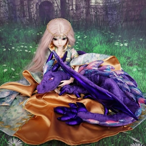 May include: A doll with long pink hair and a gold crown sits on a green grassy surface. The doll is wearing a gold and green dress and is holding a purple plush dragon.
