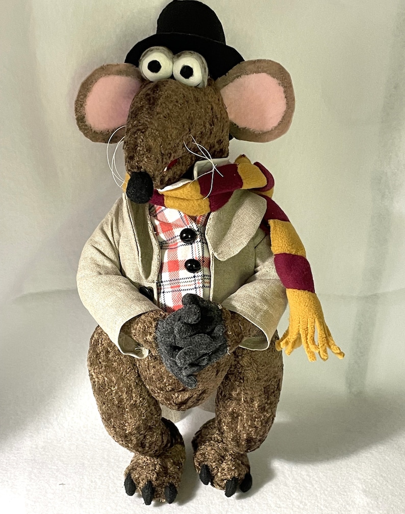 Op de afbeelding: Een bruine pluche rat met een zwarte hoed, een beige jas, een rood-wit geruit overhemd en een geel-marronkleurige sjaal. De rat zit met gekruiste poten.