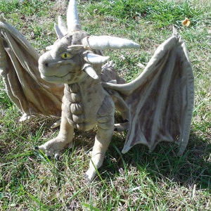 Puede incluir: Un dragón de peluche gris con ojos amarillos y alas grandes. El dragón está sentado en un lecho de hierba verde.