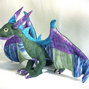 Op de afbeelding: Een groene, paarse en blauwe patchwork-stofdraak met een lange staart en vleugels. De draak heeft een geel oog en een paarse hoorn.