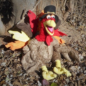 Op de afbeelding: Een pluche kalkoenpop met een pelgrimhoed en een rood, oranje en bruin verenkleed. De pop zit op een bed van bruine bladeren.