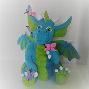 Op de afbeelding: Een blauwe en groene pluche draak met witte hoorns en vleugels. De draak houdt roze en paarse vlinders in zijn poten.