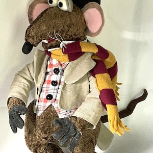 Op de afbeelding: Een bruine pluche rat die een zwarte hoed, een beige jas, een rood-gele gestreepte sjaal en een geruit vest draagt. De rat heeft grote, zwarte ogen en een lange, bruine staart.