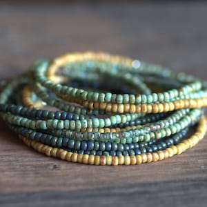 Könnte beinhalten: Ein Stapel Perlenarmbänder in Gelb-, Grün- und Blautönen. Die Armbänder bestehen aus kleinen, runden Perlen und sind in einem geschichteten Muster angeordnet.