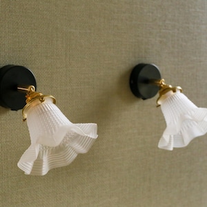 Puede incluir: Dos apliques de pared con bases circulares negras, brazos dorados y pantallas de vidrio blanco con volantes. Las pantallas tienen un patrón texturizado y acanalado. Los apliques están montados en una pared verde oliva texturizada.