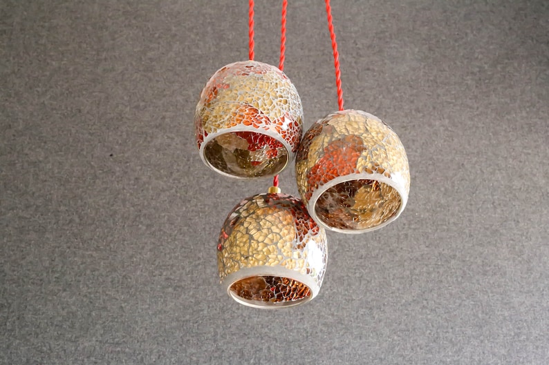May include: Three gold and red mosaic glass pendant lights hanging from red cords against a gray background.